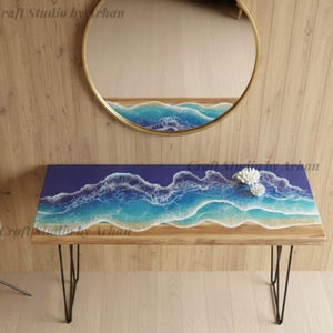 Puede incluir: Una mesa de centro de madera con un diseño de olas oceánicas azules y blancas en la parte superior, sostenida por patas de horquilla negras. Un espejo redondo dorado cuelga de la pared sobre la mesa, reflejando el diseño oceánico. Un coral blanco decora la mesa.