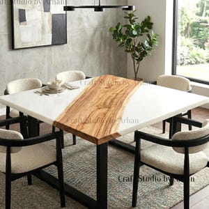 May include: A contemporary dining table featuring a white epoxy resin surface with a natural wood inlay. The table is supported by black metal legs and surrounded by four upholstered chairs. The table measures approximately 127 cm square.