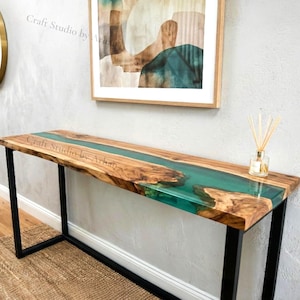 May include: A wooden console table with a teal resin river feature. The table has a natural wood grain finish and black metal legs. The wall is light grey, with a framed artwork and a circular mirror. The table is approximately 150cm long.