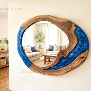 Op de afbeelding: Een ovale wandspiegel met een natuurlijke houten lijst en een levendige blauwe harsinleg. De spiegel reflecteert een woonkamerscène. De tekst "Craft Studio by Arhan" is zichtbaar.