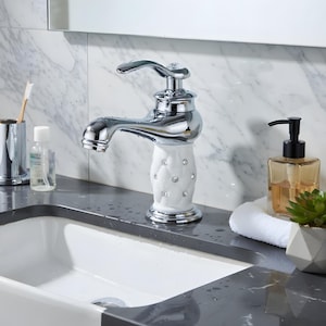 May include: A polished chrome bathroom tap with a white, rhinestone-studded base. The tap is set above a white sink, with a soap dispenser, toothbrush holder, and other bathroom accessories in the background.