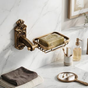 May include: Antique brass soap dish with a decorative crown design. The soap dish is mounted on a wall and holds a green bar of soap. The design includes a pivoting arm and a rectangular soap holder.
