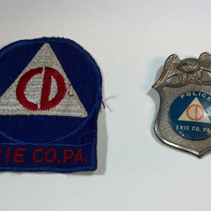 May include: Two vintage Erie County, PA items: a blue and white patch and a silver police badge. The patch features a white triangle with a red circle and the letters "CD". The badge has a blue circle with the same design and the word "POLICE".