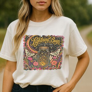 May include: Cream-colored t-shirt featuring a colorful graphic design. The design includes the text "Lightning Bugs & Lazy Summer Nights" above a jar filled with glowing lights, surrounded by flowers and butterflies.
