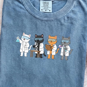 May include: A blue short-sleeved t-shirt featuring a graphic of four cartoon cats dressed as scientists in white lab coats and safety goggles. The cats are performing experiments with a microscope, clipboard, and beakers. The shirt has a Comfort Colors tag.