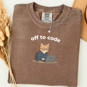 May include: Brown t-shirt with the text "off to code" and a cartoon cat wearing glasses and a hoodie, working on a laptop. A silver necklace with a star pendant is draped over the shirt.