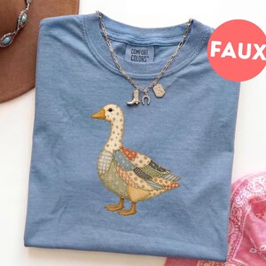 Puede incluir: Una camiseta azul claro con un gráfico central de un ganso con un patrón estilo patchwork en tonos apagados de beige, azul y naranja. La camiseta tiene una etiqueta Comfort Colors en el cuello y se combina con un collar de plata con dijes.