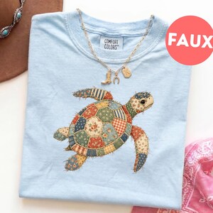 Puede incluir: Una camiseta azul claro con el gráfico de una tortuga marina cuyo caparazón está compuesto por varios patrones de tela floral y de cuadros multicolores. La camiseta tiene una etiqueta Comfort Colors en el cuello y se combina con un collar de oro.