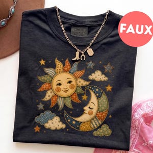Puede incluir: Una camiseta negra con un estampado gráfico de un sol sonriente y una luna creciente, ambos decorados con coloridos patrones de patchwork. El diseño incluye pequeñas estrellas y nubes. Un collar de cadena dorada con un dije de bota descansa encima.