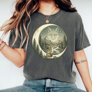 May include: Dark grey t-shirt featuring a nature-themed graphic. The design includes a fox, mountains, trees, and a river, all set within a crescent moon shape. The fox is depicted in a detailed, artistic style.