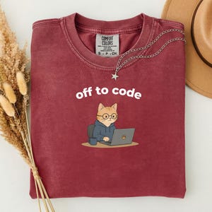 以下が含まれることがあります： 「off to code」の白い文字が入ったマルーン色のTシャツ。メガネとパーカーを着てノートパソコンで作業をしている漫画の猫が描かれています。星のペンダントが付いたシルバーのネックレスがシャツにかけられています。