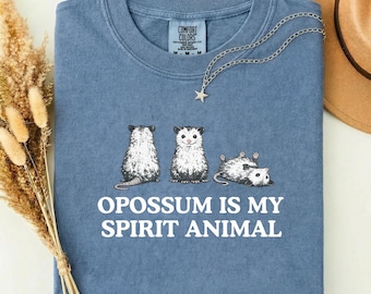 以下が含まれることがあります： 「OPOSSUM IS MY SPIRIT ANIMAL」の文字とオポッサムのグラフィックがプリントされた、青色のComfort Colors Tシャツ。星のペンダントが付いたシルバーのネックレスがシャツの上にあります。