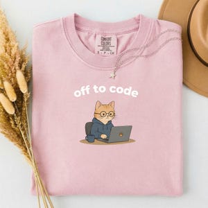 以下が含まれることがあります： 「off to code」の文字と、メガネとパーカーを着た猫のイラストがプリントされた、薄いピンク色のTシャツ。クルーネックで半袖です。