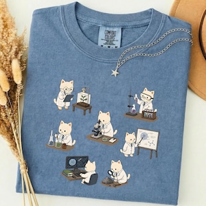 Puede incluir: Camiseta azul Comfort Colors con un gráfico de temática científica con gatos de dibujos animados con batas de laboratorio. Los gatos están representados realizando experimentos con vasos de precipitados, microscopios y un ordenador. Un collar plateado con un colgante de estrella está drapeado sobre la camiseta.