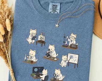 以下が含まれることがあります： 青色のComfort Colors Tシャツ。科学をテーマにしたグラフィックで、白衣を着た漫画の猫が描かれています。猫はビーカー、顕微鏡、コンピューターを使って実験を行っています。星のペンダントが付いたシルバーのネックレスがTシャツにかけられています。
