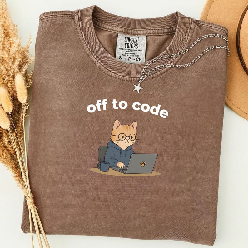 以下が含まれることがあります： 「off to code」の文字と、メガネとパーカーを着てノートパソコンで作業をしている漫画の猫が描かれた茶色のTシャツ。星のペンダントが付いたシルバーのネックレスがシャツの上にかけられています。