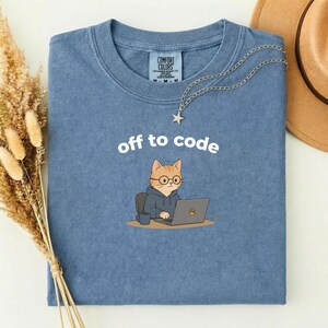 以下が含まれることがあります： 「off to code」の文字が書かれた青いTシャツ。メガネとフード付きパーカーを着た漫画の猫がノートパソコンで作業しています。星のペンダントが付いたシルバーのネックレスがシャツの上にかけられています。