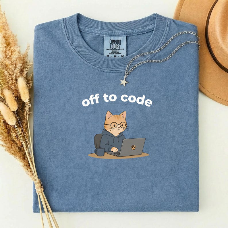 以下が含まれることがあります： 「off to code」の文字が書かれた青いTシャツ。メガネとフード付きパーカーを着た漫画の猫がノートパソコンで作業しています。星のペンダントが付いたシルバーのネックレスがシャツの上にかけられています。