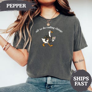 Puede incluir: Una camiseta de manga corta gris carbón con un gráfico de un ganso blanco con gafas y sudadera con capucha, que lleva una mochila y un portátil. El texto sobre el ganso dice: "off to do coding things". La camiseta tiene cuello redondo.