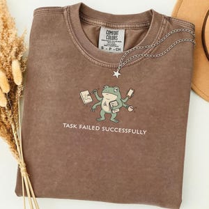 Puede incluir: Una camiseta Comfort Colors marrón con un dibujo de una rana. La rana sostiene un bolígrafo, un teléfono, un bloc de notas y una taza de café. El texto "TASK FAILED SUCCESSFULLY" está debajo. Una cadena plateada con un colgante de estrella está en la camiseta.
