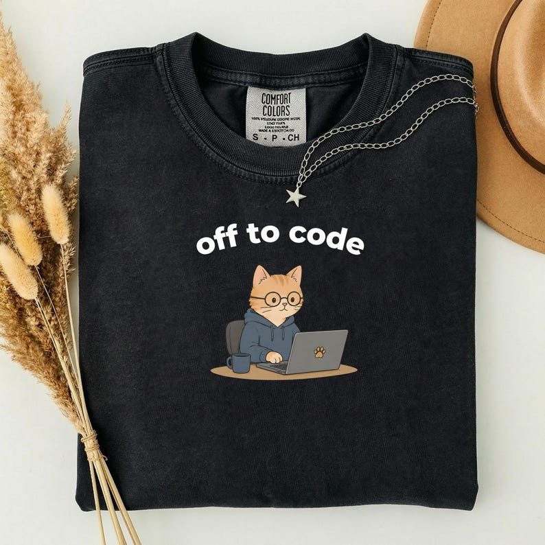 以下が含まれることがあります： 「off to code」の文字と、メガネとパーカーを着てノートパソコンで作業をしている漫画の猫が描かれた黒いTシャツ。星のペンダントが付いたシルバーのネックレスがシャツにかけられています。シャツにはComfort Colorsのタグが付いています。