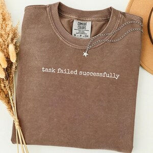 Puede incluir: Una camiseta Comfort Colors marrón con la frase "task failed successfully" impresa en blanco. Un collar de cadena plateada con un colgante de estrella está colocado sobre la camiseta. Una parte de un sombrero marrón es visible en el fondo.