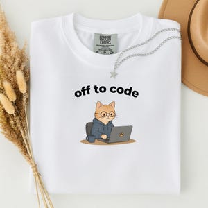 以下が含まれることがあります： 「off to code」の文字と、メガネとパーカーを着てノートパソコンで作業をしている漫画の猫が描かれた白いTシャツ。猫はコーヒーカップと一緒に椅子に座っています。