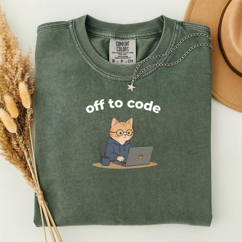 以下が含まれることがあります： 「off to code」の文字がプリントされたオリーブグリーンのTシャツ。メガネとフード付きパーカーを着た漫画の猫がノートパソコンで作業しています。星のペンダントが付いたシルバーのネックレスがシャツの上にあります。