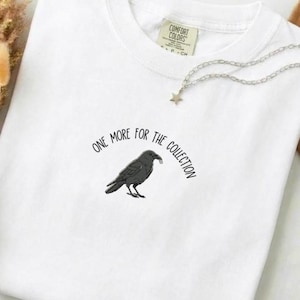 Puede incluir: Camiseta blanca con un gráfico de un cuervo negro sosteniendo un pequeño objeto en su pico. El texto "ONE MORE FOR THE COLLECTION" está arqueado alrededor del cuervo. Un collar plateado con un colgante de estrella está sobre la camiseta.