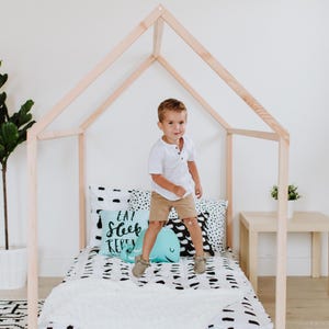 May include: A young boy jumps on a bed with a wooden frame shaped like a house. The bed has a white and black patterned sheet and a pillow with the text "Eat Sleep Repeat" in black lettering.