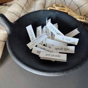 May include: A black ceramic bowl filled with small white slips of paper, each with a grade level written on it, including preschool, pre-k, 1st grade, 3rd grade, 7th grade, 9th grade, 10th grade, and 11th grade.