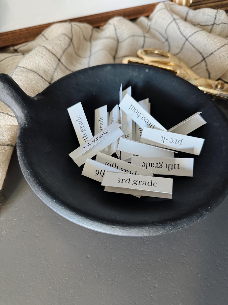 May include: A black ceramic bowl filled with small white slips of paper, each with a grade level written on it, including preschool, pre-k, 1st grade, 3rd grade, 7th grade, 9th grade, 10th grade, and 11th grade.