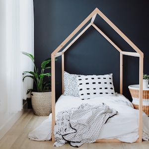 May include: A white bed with a black and white patterned blanket and pillows inside a wooden house-shaped frame. The bed is on a wooden floor in a room with a white wall and a plant in a basket.