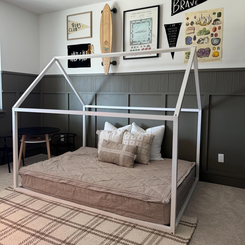 May include: A white wooden bed frame with a house-shaped canopy. The bed has a brown mattress and a few pillows on top. The bed is in a room with gray walls and a rug on the floor.