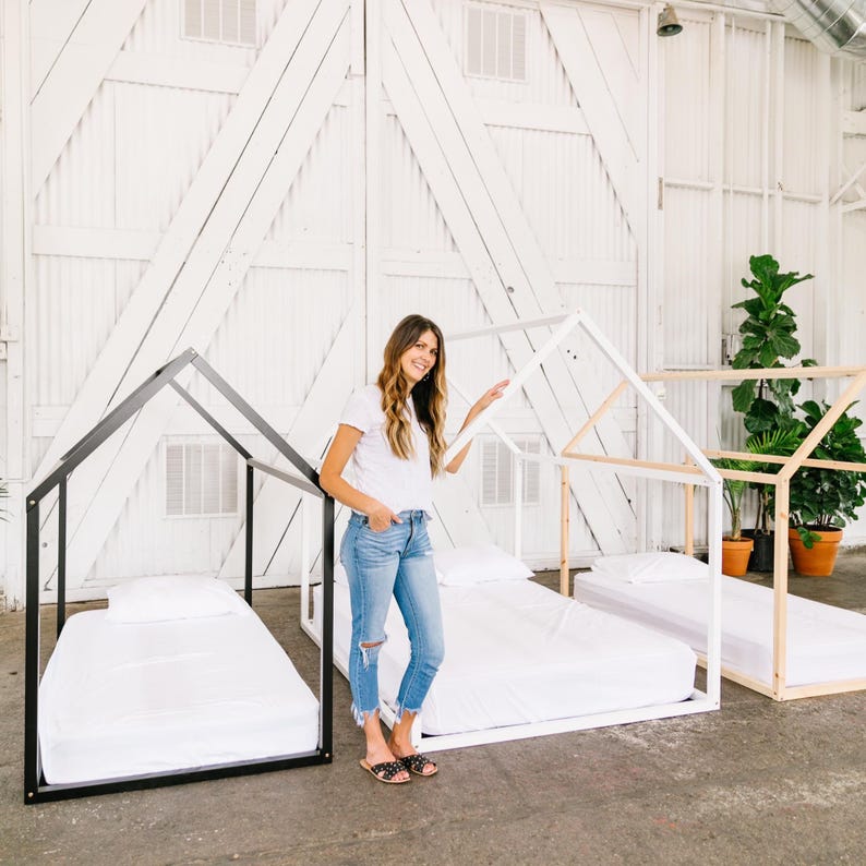 May include: Three white and black wooden bed frames in the shape of a house. The bed frames are set up in a room with white walls and a white door. A woman is standing in front of the bed frames.