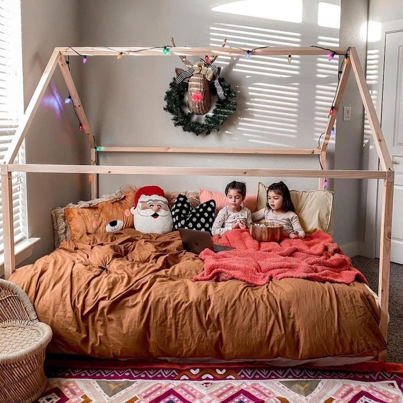 May include: Two children are sitting in a wooden bed frame decorated with Christmas lights. They are both wearing red shirts and are looking at each other. The bed has a brown comforter and a red blanket. There are pillows on the bed, including a Santa Claus pillow.