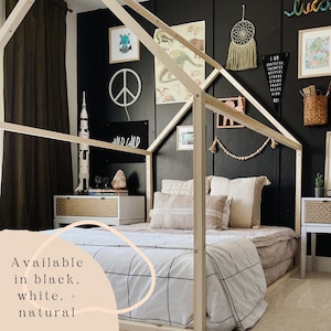 May include: A wooden house-shaped bed frame with a white and black checkered duvet and pillows. The bed is in a room with black walls and a variety of decorations, including a peace sign, a rocket ship, and a dream catcher. The text "Available in black, white, + natural" is visible in the lower left corner of the image.
