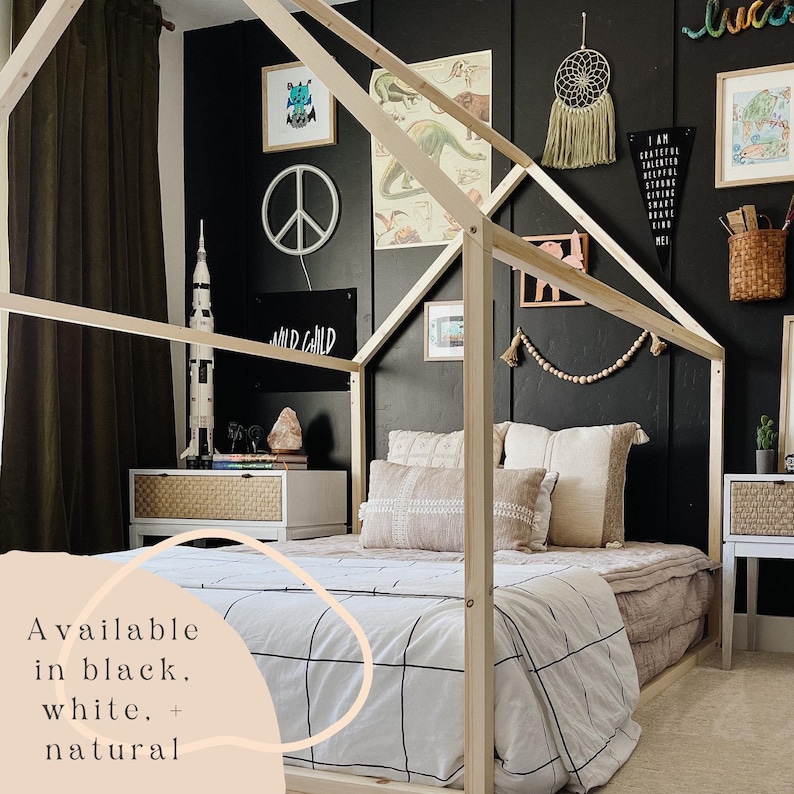 May include: A wooden house-shaped bed frame with a white and black checkered duvet and pillows. The bed is in a room with black walls and a variety of decorations, including a peace sign, a rocket ship, and a dream catcher. The text "Available in black, white, + natural" is visible in the lower left corner of the image.