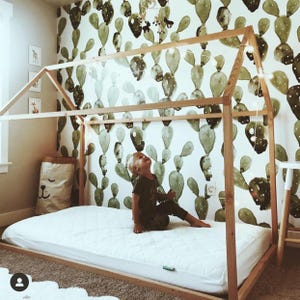 May include: A wooden bed frame in the shape of a house with a white mattress and a child sitting on the bed. The wall behind the bed is covered in green cactus wallpaper.