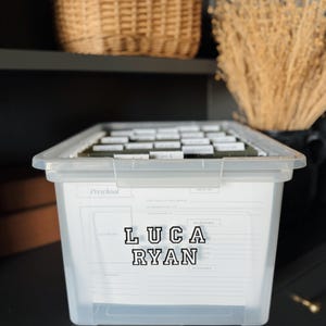 May include: A clear plastic storage bin with a white label that says "Preschool" and has the name "Luca Ryan" written in black letters. The bin is filled with labeled file folders.