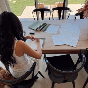 May include: A person sits at a wooden table with a stack of green folders and several blank white sheets of paper. The person is writing on one of the sheets of paper. There is a dog sitting on the floor next to the person.