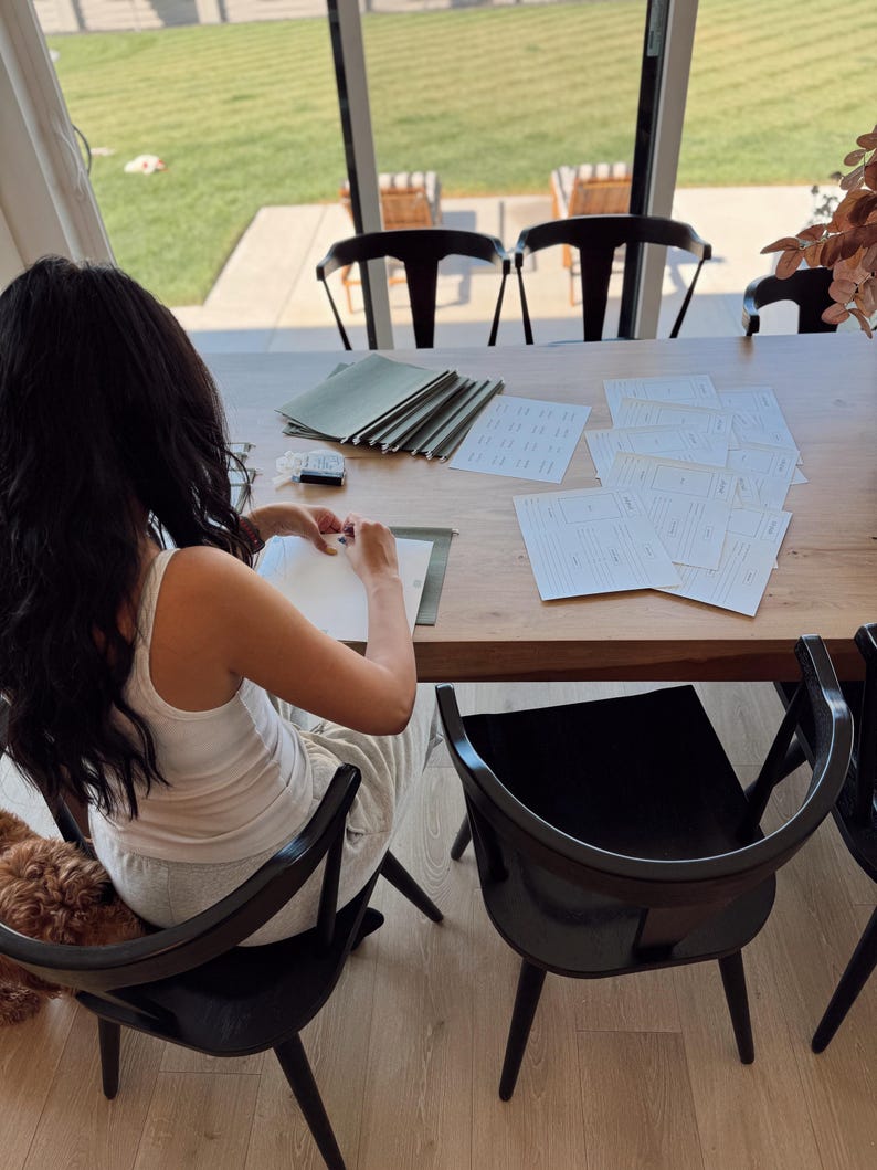 May include: A person sits at a wooden table with a stack of green folders and several blank white sheets of paper. The person is writing on one of the sheets of paper. There is a dog sitting on the floor next to the person.