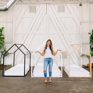 May include: Two wooden bed frames with white mattresses, one black and one natural wood, in a white room with a woman standing in the middle.