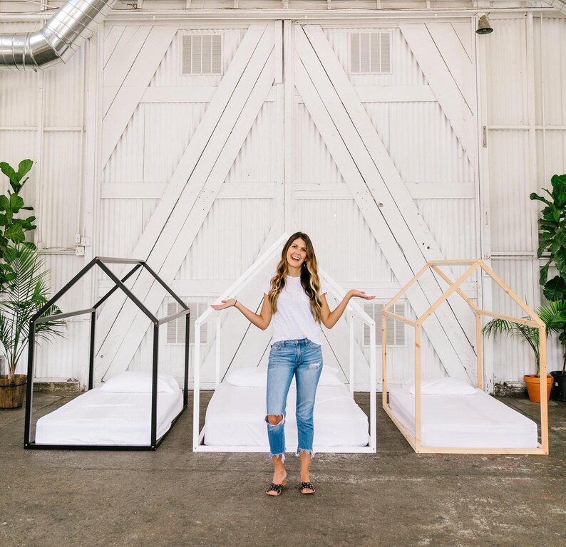 May include: Two wooden bed frames with white mattresses, one black and one natural wood, in a white room with a woman standing in the middle.