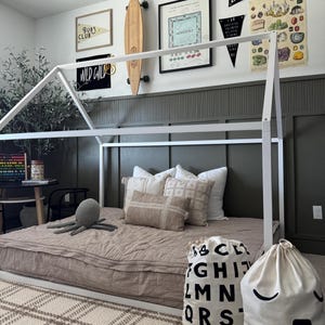 May include: A white wooden playhouse bed frame with a brown quilted mattress. The bed is decorated with throw pillows and two white canvas bags with black lettering. One bag has the alphabet printed on it and the other has a simple smiley face.
