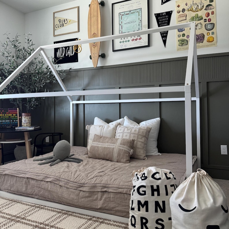May include: A white wooden playhouse bed frame with a brown quilted mattress. The bed is decorated with throw pillows and two white canvas bags with black lettering. One bag has the alphabet printed on it and the other has a simple smiley face.