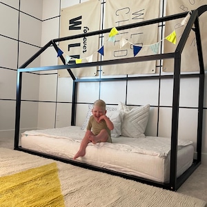 May include: A black metal frame bed with a white mattress and a child sitting on it. The bed is decorated with a yellow and blue bunting. The wall behind the bed has a white tile pattern.