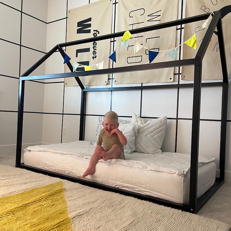 May include: A black metal frame bed with a white mattress and a child sitting on it. The bed is decorated with a yellow and blue bunting. The wall behind the bed has a white tile pattern.