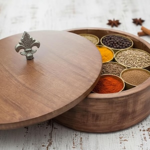 Op de afbeelding: Een houten kruidenbox met een deksel en een decoratieve metalen handgreep. De box is open en toont kleine metalen bakjes gevuld met diverse kruiden, waaronder kurkuma, komijn en rode peper. Op een lichtgekleurd houten oppervlak.