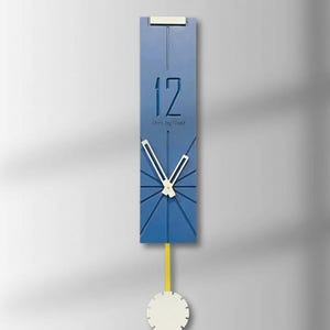 Könnte beinhalten: Eine moderne, blaue Wanduhr mit weißem Pendel und Zeigern. Das Zifferblatt zeigt die Zahl 12 und den Text "Urns by Naaz". Das Pendel ist gelb und die Uhr ist rechteckig.